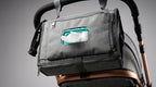 A dark gray diaper bag with a front zippered pocket holding a turquoise tissue box, shown hanging from a stroller handle on a neutral gray background.