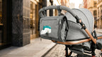 A gray diaper bag with a built-in tissue dispenser, mounted on the back of a stroller, shown in an outdoor urban setting.